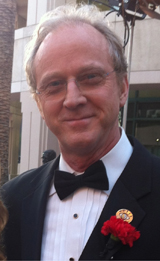 A man with light hair and glasses wearing a black tuxedo and bowtie with a red flower boutonniere.
