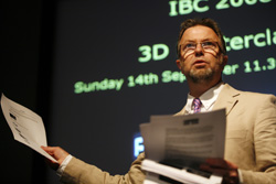 A man in a tan suit and glasses holds papers while standing before a dark screen with green text.