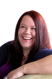 A smiling woman with long reddish-brown hair wearing a purple shirt and dark cardigan.