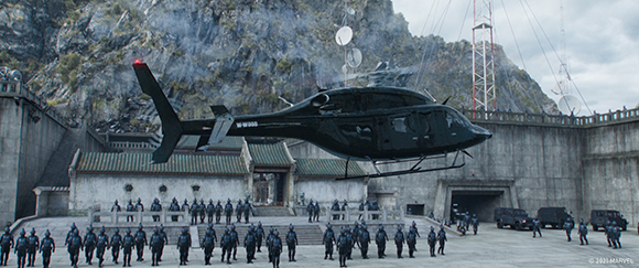 A black helicopter flies low over rows of soldiers standing in a courtyard before a stone fortress.