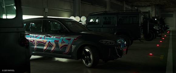 A black car with pink and blue graffiti-style text parked in a dim garage next to dark SUVs.