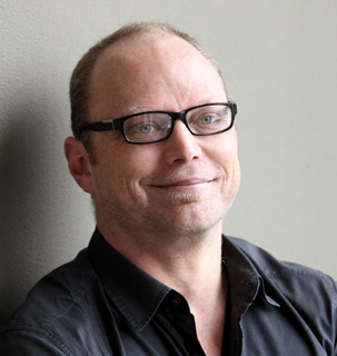 A man with short hair and glasses smiling against a plain wall.