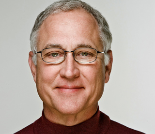 A man with short gray hair and glasses wearing a maroon turtleneck against a light background.
