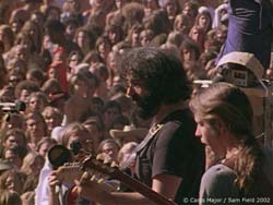 A bearded man plays a guitar facing a large, dense crowd of people at an outdoor event.
