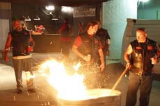 Several men stand around a large, bright fire in a dark, outdoor or industrial setting.