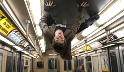 A man in a brown jacket hangs upside down from the ceiling of a subway train car.