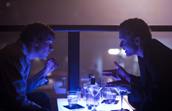 Two men sit across from each other at a small table in a dimly lit bar with blue lighting.