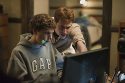 Two young men lean in to look intently at a computer screen in a dimly lit room.