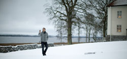 A man in a grey jacket walks through snow toward a lake with bare trees and a building in the background.