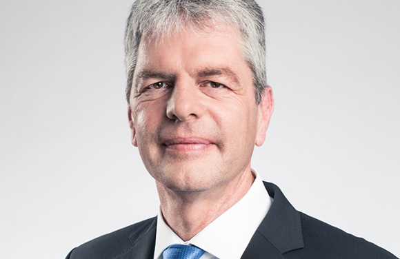 A middle-aged man with graying hair wearing a black suit, white shirt, and blue tie against a light gray background.