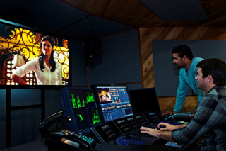 Two men monitor video production equipment in a dark studio with a large screen showing a woman.