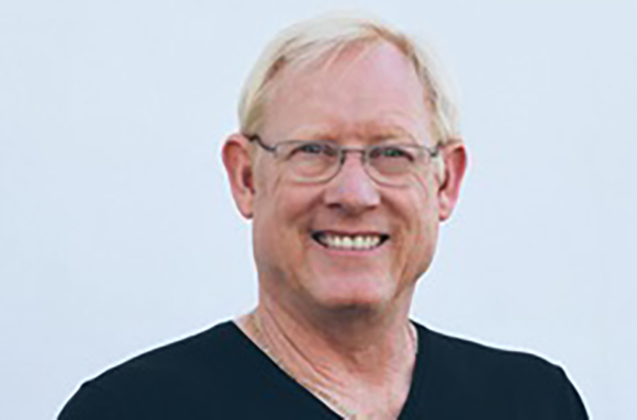 A smiling man with short blonde hair and glasses wearing a black shirt against a light background.
