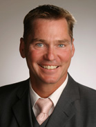 A smiling man with short brown hair wearing a dark suit, white shirt, and light pink tie.