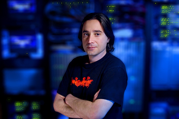 A man with dark hair and a black t-shirt stands with arms crossed in a blue-lit server room.