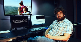 A man with dark hair and a beard sits at a desk with three computer monitors in a dark room.