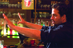 A man in a black shirt extends both arms forward with palms facing outward in a dimly lit bar.