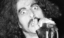 A man with long, dark curly hair and a mustache sings intensely into a microphone.
