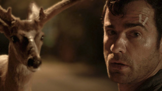 A man with a small bandage on his forehead looks forward next to a deer with antlers at night.