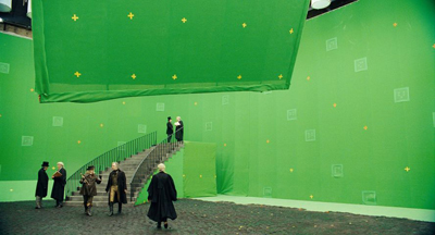 Actors in period costumes walk near a staircase on a large green screen film set.