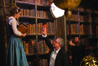 A woman in a blue dress holds a book as a man in a black jacket gestures toward a large library bookshelf.