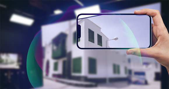 A hand holds a smartphone horizontally to take a photo of a white building with green window shutters.