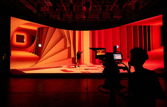 A silhouette of a camera operator filming a person standing before a large, red-lit geometric set.
