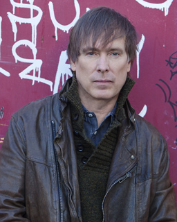 A man with light brown hair wearing a brown leather jacket and a dark green textured collar.