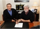 Two men sit at a dark desk with a pen and a piece of paper in an office setting.