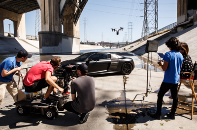 Film crew using a camera dolly and drone to film a black sports car in a concrete drainage canal.