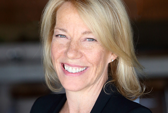 A smiling blonde woman with blue eyes wearing a black blazer.