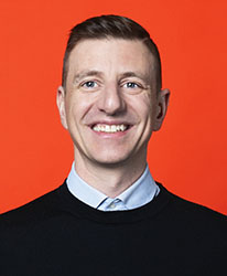 A smiling man with short brown hair wearing a light blue collared shirt and a black sweater.