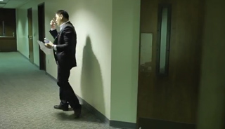A man in a dark suit walks down a long, brightly lit office hallway while holding papers.