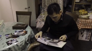 A man in a black sweater sits and looks through a large open photo album in a dimly lit room.