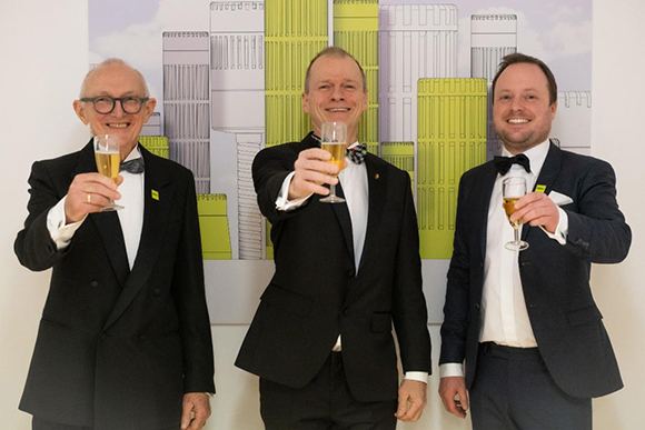 Three men in black suits and bow ties toast with champagne glasses against a graphic background.