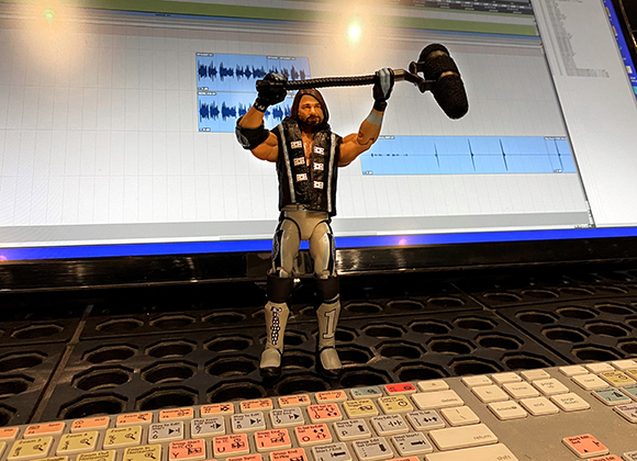 An action figure in a vest holds a large microphone hammer up against a computer monitor displaying audio waves.