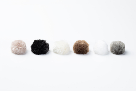 Six fuzzy pom-poms in pink, black, cream, brown, white, and grey arranged in a horizontal line on a white background.