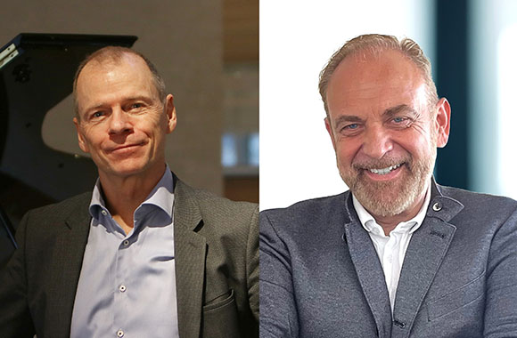 Two smiling men in business casual attire, one in a light blue shirt and one in a white shirt and grey blazer.