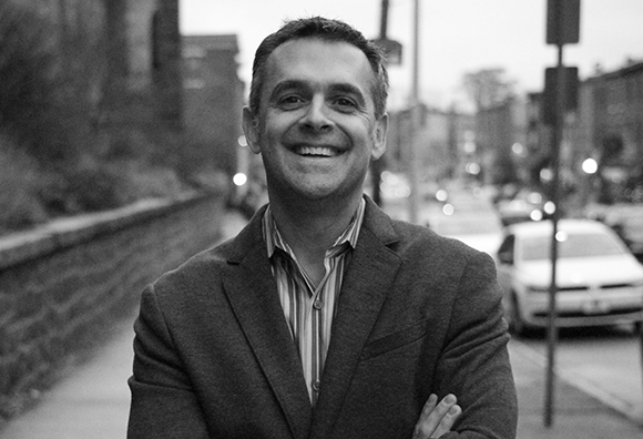 A smiling man in a blazer and striped shirt stands on a city street in this black and white portrait.