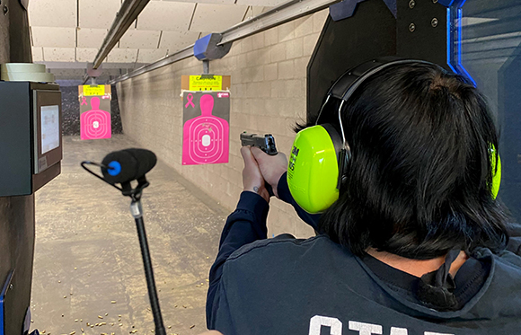 A person with black hair wearing neon green earmuffs aims a handgun at pink target silhouettes.