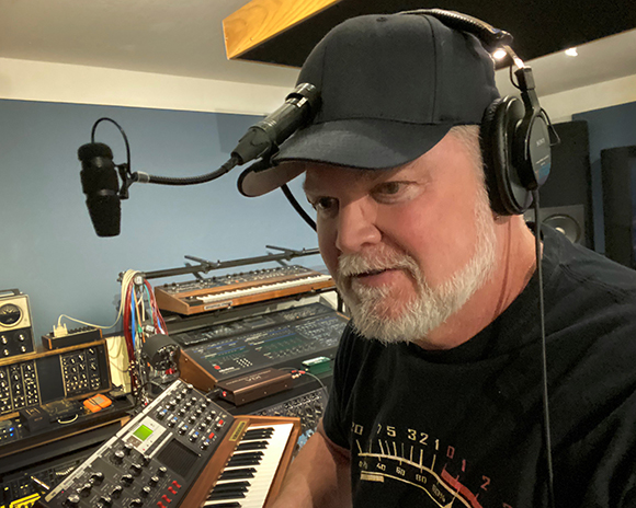 A man with a grey beard wearing a black cap and headphones in a room filled with synthesizers.