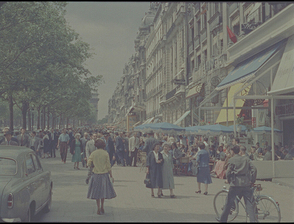 Pedestrians walk along a wide city sidewalk lined with multi-story buildings and outdoor cafe awnings.