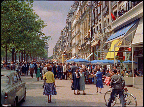 People walk along a wide sidewalk past outdoor cafe seating and tall European-style buildings.