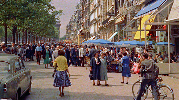 People walking and dining at outdoor cafes along a tree-lined city boulevard with tall buildings.