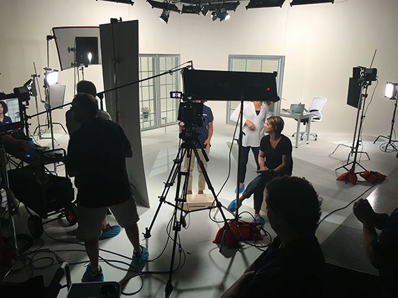 A film crew uses cameras and lighting equipment to film two women in a white studio set.