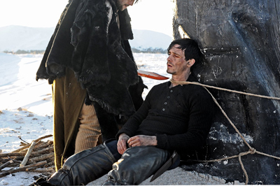 A man in a black shirt is tied to a dark tree trunk with rope while another person stands beside him.