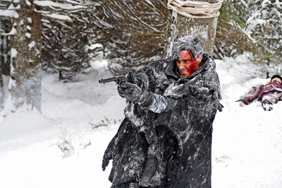 A man with blood on his face aims a handgun while wearing a dark, snow-covered coat in a snowy forest.