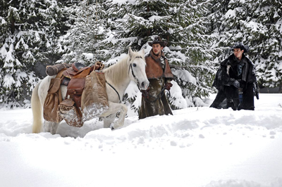 A man in a brown coat stands beside a white horse laden with gear in a snowy forest.