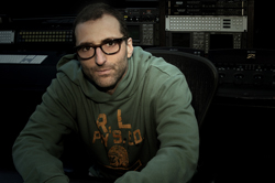 A man with glasses wearing a green hoodie sits in a dark recording studio with audio equipment behind him.