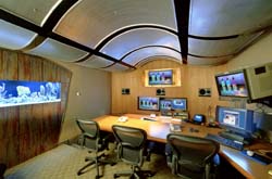 A control room featuring a curved paneled ceiling, a long desk with multiple computer monitors, and black office chairs.