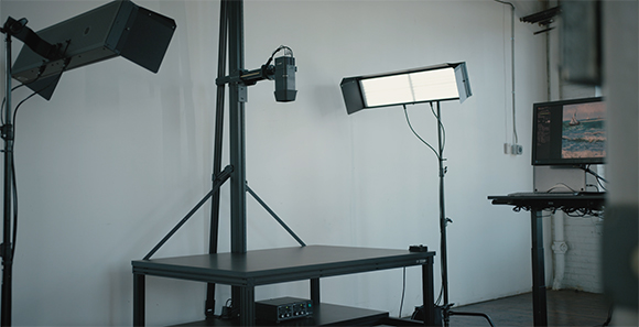 A photography studio featuring softbox lights, a camera on a stand, and a monitor on a desk against a white wall.